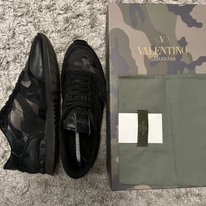 Valentino Rockrunner sneakers camouflage - Snygga Valentino Rockrunner sneakers med mörkt kamouflagemönster i svart, grått och grönt. Endast skorna medföljer. Skorna har platt sula med nitar på sidan, snörning och detaljer i mocka och skinn. Perfekt för dig som vill ha en cool och exklusiv look.