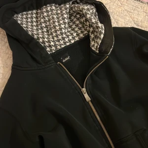 Svart vindjacka med mönstrad huva - Svart zip-hoodie från deVal med dragkedja framtill och två fickor med metallöglor. Insidan av huvan har ett snyggt svartvitt rutigt mönster. Jackan har lång ärm och en clean, sportig look som funkar till streetwear. Har fått som gelé vid en av ärmarna (skriv gärna om ni vill ha bild på det)☺️