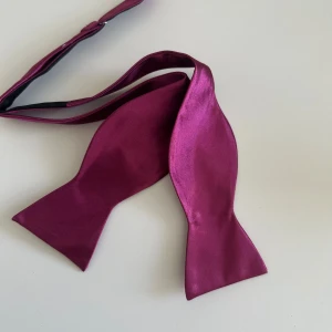 Vinröd självknuten fluga i satin - Självknuten fluga i vinröd satin med justerbart band. Snygg och stilren accessoar med glansig finish som ger en lyxig känsla. Perfekt för att lyfta din kostym till nästa nivå. Flugan levereras i plastförpackning.