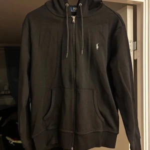 Svart hoodie från Polo Ralph Lauren - Svart hoodie från Polo Ralph Lauren med klassisk broderad logga på bröstet. Tröjan har huva med snörning, dragkedja framtill och två rymliga fickor. Tillverkad i mjuk bomull, perfekt för chill dagar. Stilren och enkel design som alltid funkar.