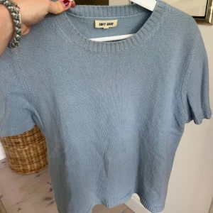 Ljusblå stickad tröja från Soft Goat - Superfin ljusblå stickad tröja från Soft Goat med korta ärmar och rund halsringning. Tröjan är i mjuk kashmir och har ribbade detaljer vid hals, ärmslut och nederkant. Perfekt för lager-på-lager eller sommarens svalare dagar. Den har några små fläckar men man tänker inte på dem!