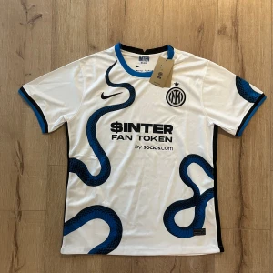 Inter Milan vit fotbollströja Nike M - Säljer en officiell Inter Milan fotbollströja från Nike i storlek M. Tröjan är vit med svarta och blå detaljer, har ett unikt ormmönster och tryck på bröstet och ryggen. Rund hals med blå kant, korta ärmar och tillverkad i Dri-FIT polyester för bästa komfort.