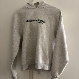 Grå hoodie från Gina Tricot - Mysig ljusgrå hoodie från Gina Tricot med texten 'Manhattan Valley New York' broderad framtill. Tröjan har huva, långa ärmar och ribbade muddar🥰