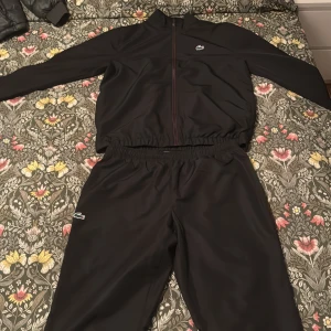 Svart träningsset från Lacoste - Säljer ett svart träningsset från Lacoste med jacka och byxor. Jackan har dragkedja och stor broderad krokodil på ryggen, samt liten logga på bröstet. Byxorna har resår i midjan och liten krokodillogga på benet. Perfekt för sport eller chill.