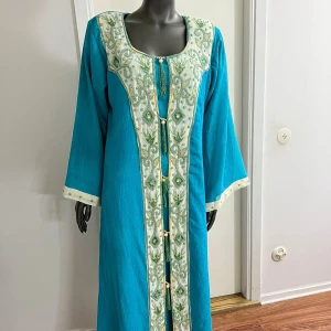 Turkos långklänning med broderi - Unik turkos långklänning med breda ärmar och vackra broderade detaljer i grönt och guld längs halsringning och framtill. Klänningen har dekorativa pärlor, tofsar och ett elegant mönster som ger en lyxig känsla. Perfekt för dig som vill sticka ut med stil.