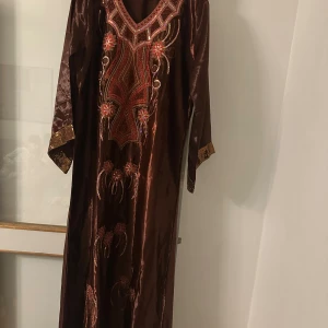 Brun långklänning med broderi och paljetter - Snygg brun långklänning i glansigt material med v-ringning, långa ärmar och detaljerat broderi framtill i rött och guld. Paljetter och blommiga mönster ger en lyxig känsla. Ärmsluten har breda paljettkanter som sticker ut.