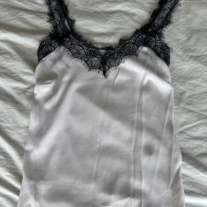 Vit satinlinnetopp med spets från M&S - Supersnygg vit linnetopp i satin från M&S med svarta spetsdetaljer längs urringningen och axelbanden. 💕passar som storlek s