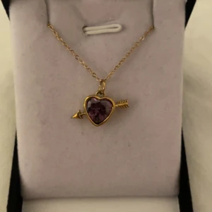 Halsband med lila hjärthänge och pil guld  - Gulligt halsband med tunn guldfärgad kedja och ett hänge i form av ett lila hjärta med en pil genom. Hjärtat är infattat i guldtonad metall och har en glittrande lila sten som ger extra wow-känsla. Perfekt accessoar för att lyfta din look. Rostfri 