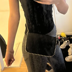 Svart axelväska med silverdetaljer - Snygg svart axelväska med silverkedja och coola nitar runt kanten. Väskan har en stilren rektangulär form och ett dekorativt hänge med dragkedja på sidan. Perfekt storlek för mobil, plånbok och nycklar.