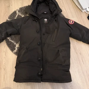 Canada Goose Chateau - Säljer en svart dunjacka från Canada Goose med huva och klassisk logga på ärmen. Jackan har dragkedja och knappar framtill, flera fickor och är perfekt för kalla dagar. Snygg och tidlös design som håller dig varm hela vintern.