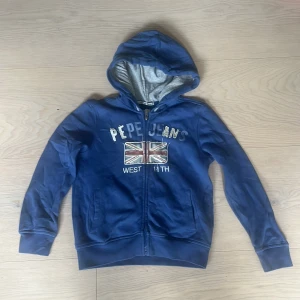 Blå hoodie från Pepe Jeans med tryck - Blå original hoodie från Pepe Jeans med Union Jack-tryck och text på bröstet. Tröjan har huva, dragkedja framtill och två fickor. Insidan av huvan är grå. Perfekt för dig som gillar streetstyle och vill ha något bekvämt och stiligt på dig. Storlek 134