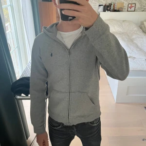 Grå hoodie från Polo Ralph Lauren - Snygg grå hoodie från Polo Ralph Lauren med dragkedja och huva. Klassisk logga broderad på bröstet, två fickor fram och mjukt bomullsmaterial. Perfekt för chill dagar och enkel att matcha med jeans eller joggers.