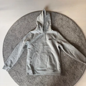Grå hoodie från Dickies med huva - Säljer en grå hoodie från Dickies med klassisk huva och snörning. Tröjan har en stor magficka och ribbade muddar vid ärmslut och nederkant. Perfekt för chill dagar och streetwear-stil.
