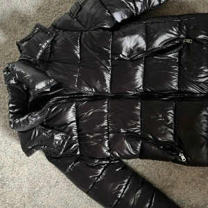 Svart glansig pufferjacka med huva - Säljer en svart pufferjacka med glansig finish och huva. Jackan har dragkedja framtill, två fickor med dragkedja och quiltad design. Perfekt för kalla dagar när du vill hålla stilen och värmen.