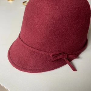 Vinröd ullhatt från Zara - Säljer en vinröd hatt från Zara i 100% ull. Hatten har en klassisk klockform med kort brätte och en smal dekorativ rosett runt kronan. Perfekt accessoar för att lyfta din look med en touch av retrostil. Snöret runt har släppt lite på ett ställe men inget som syns.