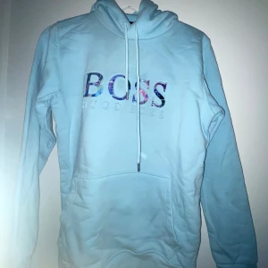 Ljusblå hoodie från Hugo Boss - Säljer en ljusblå hoodie från Hugo Boss med färgglatt BOSS-tryck på bröstet. Tröjan har huva med snörning och en stor magficka. Perfekt för dig som gillar stilrena och bekväma plagg med en twist.