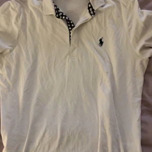 Vit Polo Golf pike från Ralph Lauren - Vit kortärmad piké från Polo Golf Ralph Lauren med svart broderad logga på bröstet, i storlek L men sitter som M. Kragen har ett snyggt rutigt mönster i svart och vitt som ger extra stilpoäng. Piken är gjord i mjuk bomull och har klassisk pikédesign. 