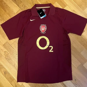 Arsenal Highbury 2006 Henry Nike tröja - Säljer en mörkröd Arsenal Highbury 1913-2006 fotbollströja från Nike med Henry 14 på ryggen. Tröjan har korta ärmar, klassisk krage och är gjord i lätt, ventilerande Dri-Fit material. Guldiga detaljer och Arsenal-emblem på bröstet. Perfekt för dig som älskar fotbollshistoria.