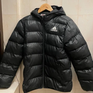 Svart pufferjacka från Adidas - Säljer en svart pufferjacka från Adidas med huva och dragkedja framtill. Jackan har Adidas-logga på bröstet och två sidofickor med dragkedja. Perfekt för kalla dagar och riktigt skön att bära. Passar dig som gillar sportig och clean stil.