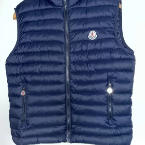 Mörkblå dunväst från Moncler - Snygg mörkblå dunväst från Moncler med quiltad design och ikonisk logga på bröstet. Västen har två fickor med dragkedja och en kraftig dragkedja framtill med Moncler-detalj. Perfekt för lager-på-lager och chill stil.