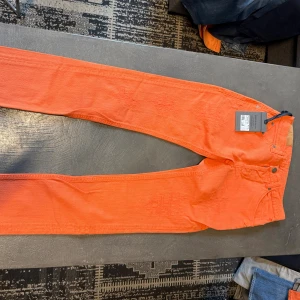 Orange jeansbyxor från Tommyville - Snygga orange jeansbyxor från Tommyville i klassisk femficksmodell. Byxorna har en rak passform och är tillverkade i bomull med lätt slitna detaljer för en cool look. Perfekt för dig som vill sticka ut med färg och stil.