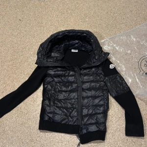 Svart Moncler pufferjacka med huva - Svart pufferjacka från Moncler med stickade ärmar och quiltad front. Jackan har en stor huva, dragkedja framtill och Moncler-logga på ärmen. Materialmix av polyester och stickad textil ger en cool kontrast. Perfekt för dig som gillar streetstyle och exklusiva märken.