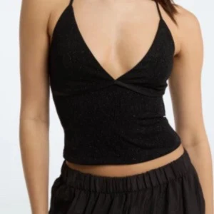 Svart glittrig croppad linnetopp - Snygg svart croppad linnetopp med tunna axelband och glittrigt material. Djup v-ringning framtill ger en trendig look, perfekt för att sticka ut. Toppen har en figurnära passform och är superenkel att matcha med både kjol och byxor.