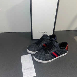 Gucci sneakers med GG-mönster - Snygga Gucci sneakers i mörkgrått med klassiskt GG-mönster, svarta skosnören och vit sula. Sidan har röd och blå rand, och baksidan har metallicröd och metallicblå detalj med Gucci-logga. Materialmix av canvas och skinn ger en lyxig känsla.