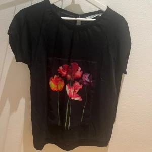 Svart blommig blus med kort ärm - Svart blus från Desigual med färgglada blommor framtill i rött, rosa och orange. Toppen har rund halsringning och korta ärmar. Materialet känns mjukt och har en lätt glansig panel framtill där blommorna är tryckta. Perfekt för dig som gillar unika prints.