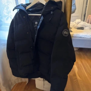 Svart pufferjacka från Canada Goose - Säljer en svart pufferjacka från Canada Goose i storlek M. Jackan har huva med snörning, dragkedja och logotyp på ärmen. Perfekt för kalla dagar med sin tjocka och quiltade design. Klassisk look och riktigt skön passform.