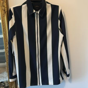 Randig skjorta med dragkedja från Zara - Snygg skjorta från Zara i relaxed fit med breda marinblå och vita ränder. Skjortan har dragkedja framtill, klassisk krage och långa ärmar. Perfekt för dig som vill sticka ut med en cool och modern look.