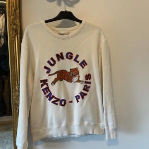 Vit Kenzo x H&M sweatshirt - säsong tog slut efter 1 månad - Vit sweatshirt från Kenzo x H&M med broderad tiger och texten 'Jungle Kenzo-Paris' i blått och rött framtill. Tröjan har rund hals, långa ärmar och ribbade muddar. Perfekt statement piece för dig som gillar streetstyle.