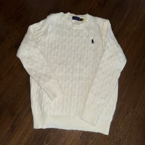 Vit kabelstickad tröja Polo Ralph Lauren - Vit kabelstickad tröja från Polo Ralph Lauren med klassisk rund halsringning och broderad marinblå logga på bröstet. Tröjan har ribbade muddar och är tillverkad i mjukt material som passar perfekt för kyligare dagar.