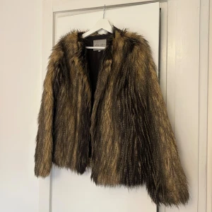 Brun pälsjacka från Ava June - Säljer en brun och svart pälsjacka från Ava June i storlek 36. Materialet är akryl och polyester. Finns  två ”hakar” så man kan stänga jackan nertill. 