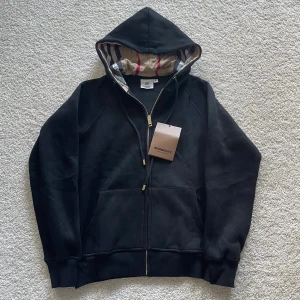 Svart Burberry hoodie med dragkedja - Svart hoodie från Burberry med klassisk rutmönstrad insida i huvan. Tröjan har guldfärgade detaljer på dragkedja och snören, samt två fickor framtill. Tillverkad i mjuk bomull och har långärmad passform med ribbade muddar. Aldrig använd! Tveka inte att höra av er vid några frågor eller funderingar🤗