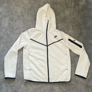 Vit Nike tech hoodie med huva - Snygg vit hoodie från Nike med svarta detaljer och dragkedja hela vägen. Hoodien har huva, lång ärm och en cool ficka med dragkedja på ena armen. Perfekt för dig som gillar sportig stil och vill ha något som sticker ut. Jag är 185 cm lång och den passar mig perfekt lite kort bara. Har bara använt den max 5 gånger