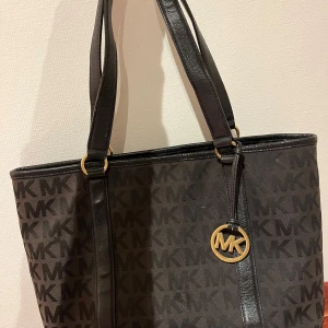 Svart axelväska från Michael Kors - Stilren svart axelväska från Michael Kors med MK-logga i mönstret och guldfärgade detaljer. Väskan har rejäla handtag, innerfack med dragkedja och magnetknäppning. Materialet är syntet med skinnliknande detaljer. Perfekt storlek för allt du behöver.