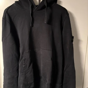 Svart hoodie från Stone Island - Svart hoodie från Stone Island med klassisk logga på ärmen. Tröjan har huva med snörning och en stor magficka framtill. Tillverkad i mjuk bomull och har en relaxed passform, perfekt för chill dagar eller streetwear-stil. Nypris 3699kr