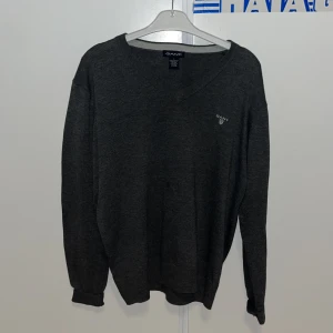 Mörkgrå stickad tröja från GANT - Snygg mörkgrå stickad tröja från GANT med v-ringad hals och diskret logga broderad på bröstet. Tröjan har långa ärmar och ribbade muddar vid ärmslut och nederkant. Perfekt för lager-på-lager under kyliga dagar.