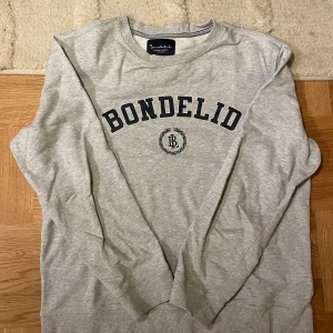 Grå sweatshirt från Bondelid - Snygg ljusgrå sweatshirt från Bondelid med klassisk logga i svart på bröstet. Tröjan har rund halsringning, långa ärmar och ribbade muddar. Perfekt för en chill och stilren look. Tillverkad i mjuk bomullsmix för extra komfort.