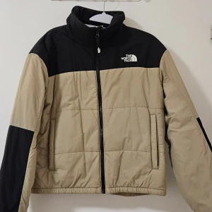 Beige och svart pufferjacka North Face - Säljer en beige och svart pufferjacka från The North Face i storlek L. Jackan har hög krage, quiltad design och dragkedja framtill. Färgblockad look med svarta partier på axlar och ärmar. Perfekt för kalla dagar och har två sidofickor.