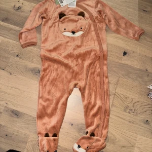 H&M varm jumpsuit, storlek 80 - Söt orangefärgad baby jumpsuit med en räv/nalle ansikte på framsidan och fötterna. Plagget har knappar hela vägen ner på framsidan är varm och skön, i nyskick! Lätt att få på och av.