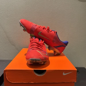 Röda Nike Mercurial fotbollsskor - Säljer ett par röda Nike Mercurial fotbollsskor med blå detaljer och grönt Swoosh-märke. Skorna har snörning, platt sula med dobbar och är tillverkade i syntetmaterial. Insidan är fodrad i blått och yttersulan har inslag av grönt och silver. Perfekta för dig som vill sticka ut på planen.