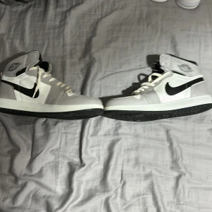 Nike Air Jordan 1 High vit/grå/svart - Säljer ett par Nike Air Jordan 1 High i vitt, grått och svart. Klassisk high-top siluett med svart swoosh, vita snören och perforerad tå. Ovandel i skinn och syntet, svart yttersula och ikoniska Jordan Wings-loggan på sidan. Perfekta för dig som vill sticka ut med streetstyle.