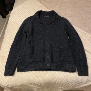 Mörkgrå stickad kofta från Southblue - Mysig mörkgrå stickad kofta från Southblue med knappar framtill och två fickor. Koftan har en klassisk krage och ribbade muddar vid ärmslut och nederkant. Perfekt för lager-på-lager under kyliga dagar.