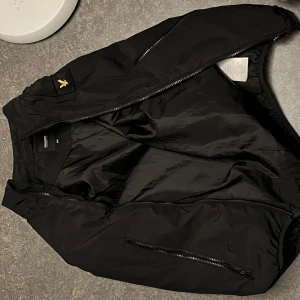 Svart bomberjacka från Lyle & Scott - Svart bomberjacka från Lyle & Scott med klassisk gul logga på axeln. Jackan har dragkedja framtill, ribbade muddar och två fickor med dragkedja. Tillverkad i polyester för en lätt och smidig känsla. Perfekt för dig som vill ha en stilren och enkel look.