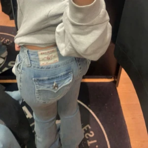 Ljusblå bootcut jeans från True religon - Säljer ett par ljusblå jeans från True religon med bootcut passform och låg midja. Jeansen har snygga detaljer som fickor med lock och metallknapp bak, samt klassisk femficksdesign.Storlek 26 väldigt stretchiga. lite söndertrampade nertill men inget som man lägger märket till vid användning. 