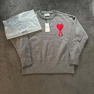 Grå stickad tröja med rött hjärta från Ami - Snygg grå stickad tröja från Ami med ett stort rött hjärta och bokstaven A broderat på bröstet. Tröjan har rund hals, ribbade muddar och nederkant samt långärmad passform. Perfekt för dig som gillar stilrena detaljer och vill sticka ut med en cool logga.