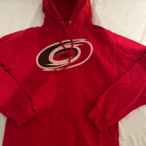 Röd NHL hoodie Carolina Hurricanes - Röd hoodie med Carolina Hurricanes-logga på bröstet och Aho 20 tryckt på ryggen. Tröjan har huva med snörning och stor magficka framtill. Materialet är mjuk bomull och passformen är normal. Perfekt för hockeyfans som vill visa sitt stöd för laget.