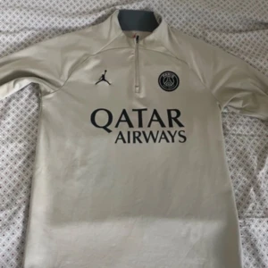 Beige PSG x Jordan långärmad tröja - Slim fit. Lite nopprig vid ärmslut annars inga defekter. Snygg beige fotbollströja från PSG och Jordan med svart logga och Qatar Airways-tryck på bröstet. Långärmad modell med halv dragkedja och ribbad krage. Tillverkad i polyester och elastan för skön passform. Perfekt för dig som gillar streetwear och fotboll.